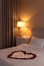 Photo of Heart made with beautiful rose petals and burning candles on bed indoors