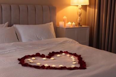 Photo of Heart made with beautiful rose petals and burning candles on bed indoors