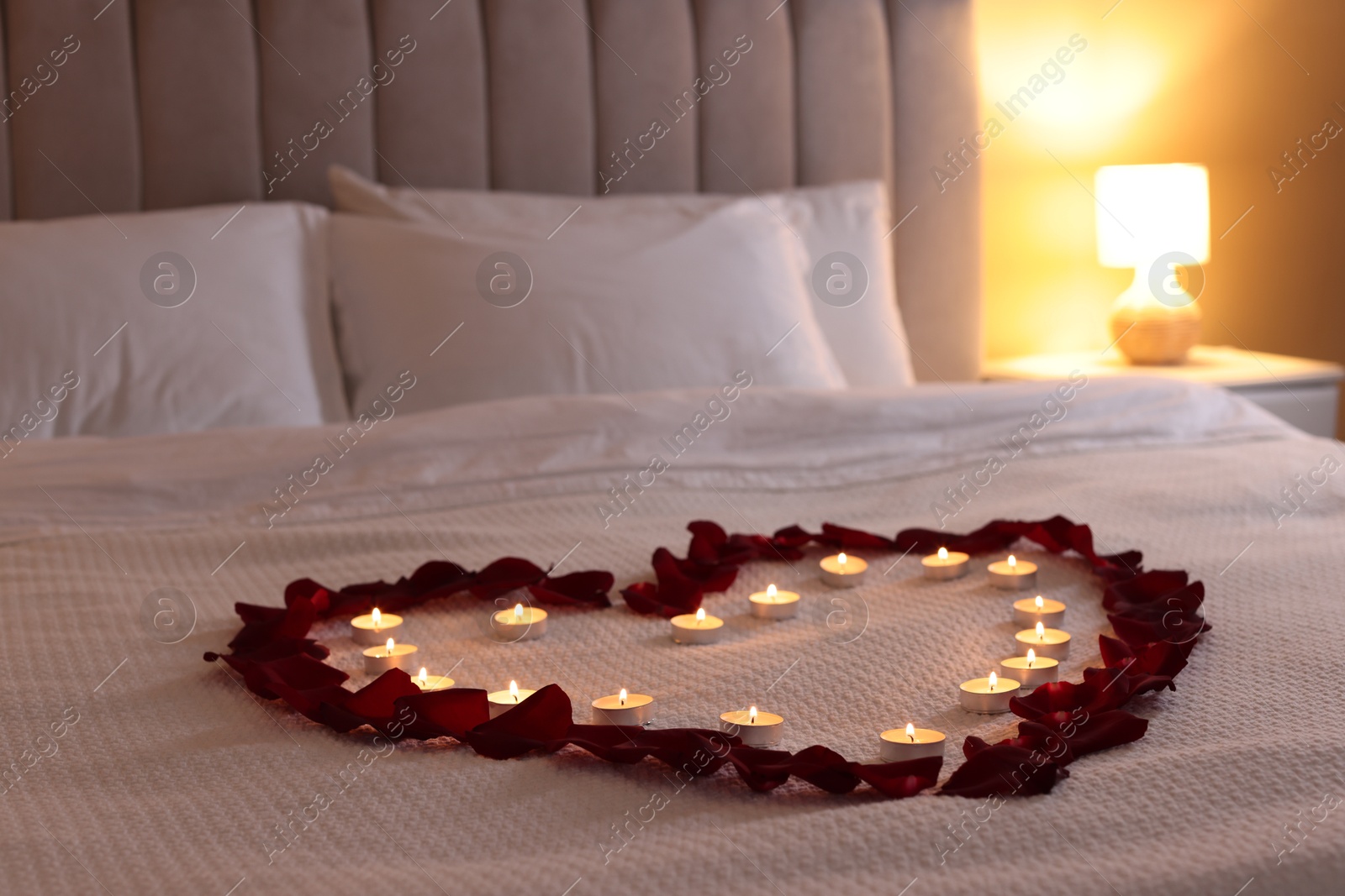 Heart made with beautiful rose petals and burning candles on bed indoors Photo of Heart made with beautiful rose petals and burning candles on bed indoors