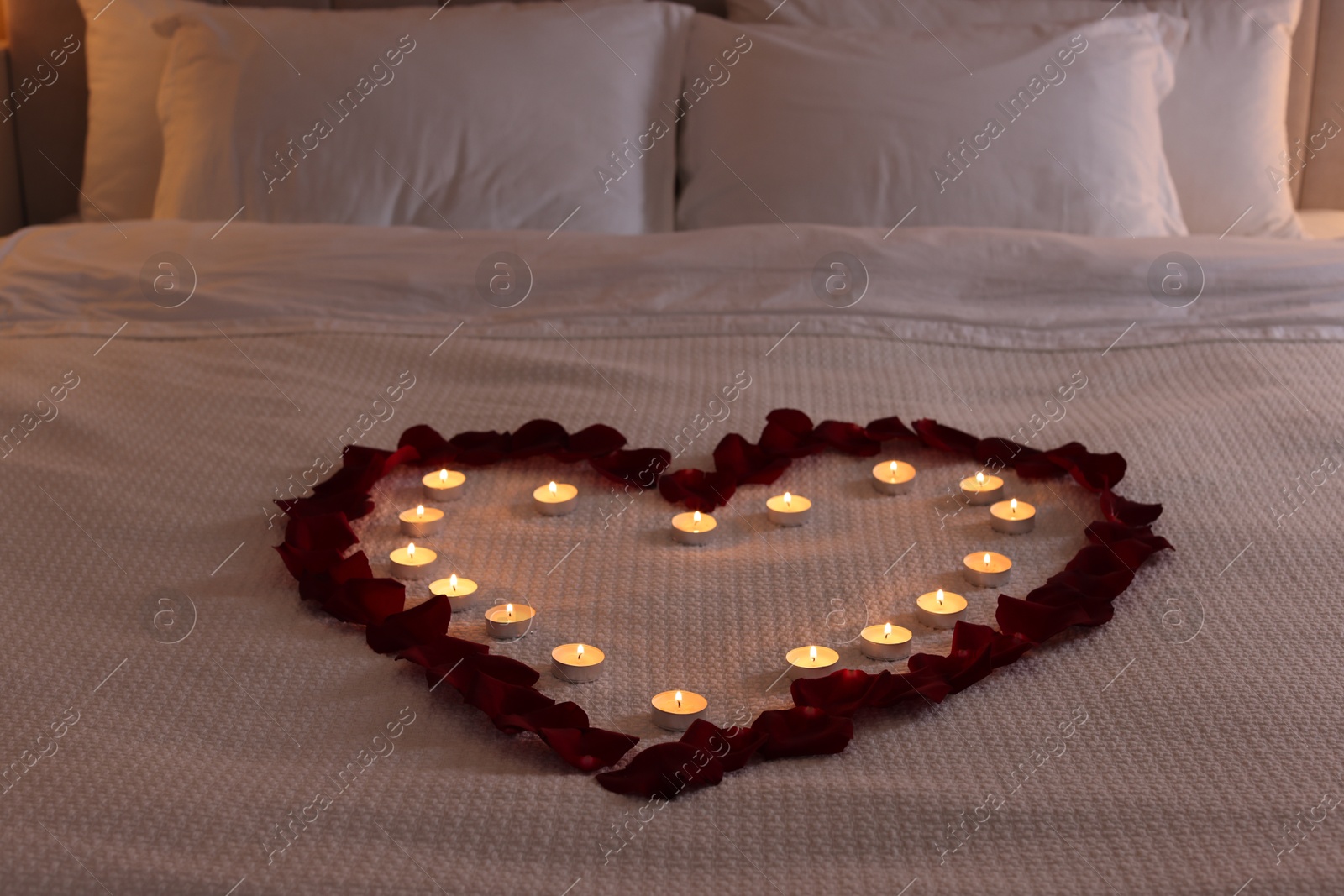 Heart made with beautiful rose petals and burning candles on bed Photo of Heart made with beautiful rose petals and burning candles on bed