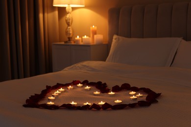 Photo of Heart made with beautiful rose petals and burning candles on bed indoors