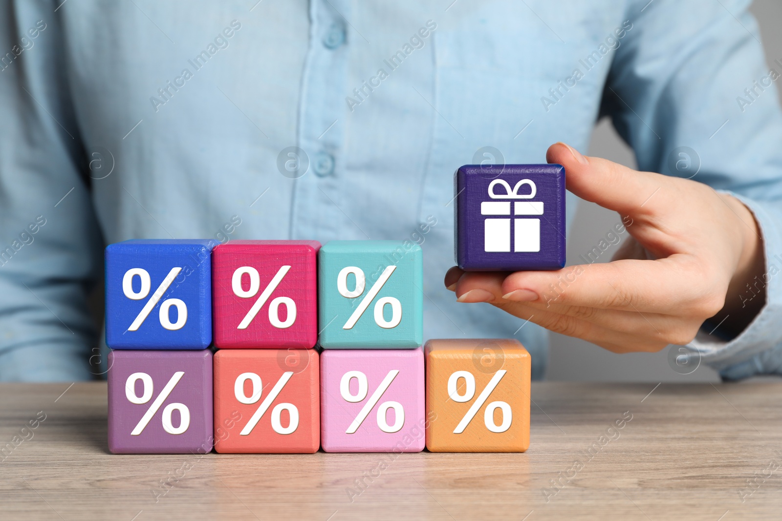 Image of Bonus. Woman holding cube with gift box image among other ones with percent signs at wooden table, closeup