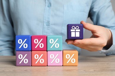 Image of Bonus. Woman holding cube with gift box image among other ones with percent signs at wooden table, closeup