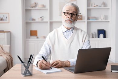 Photo of Senior man taking notes while learning online at desk indoors. Self-study