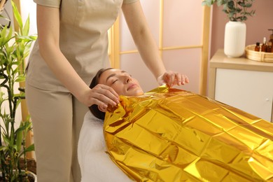 Photo of Spa body wraps. Esthetician covering woman with thermal foil blanket indoors, closeup