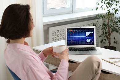 Photo of Stock exchange. Woman with cup of drink analysing financial market on laptop at white table indoors