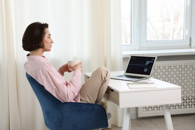 Photo of Stock exchange. Woman with cup of drink analysing financial market on laptop at white table indoors