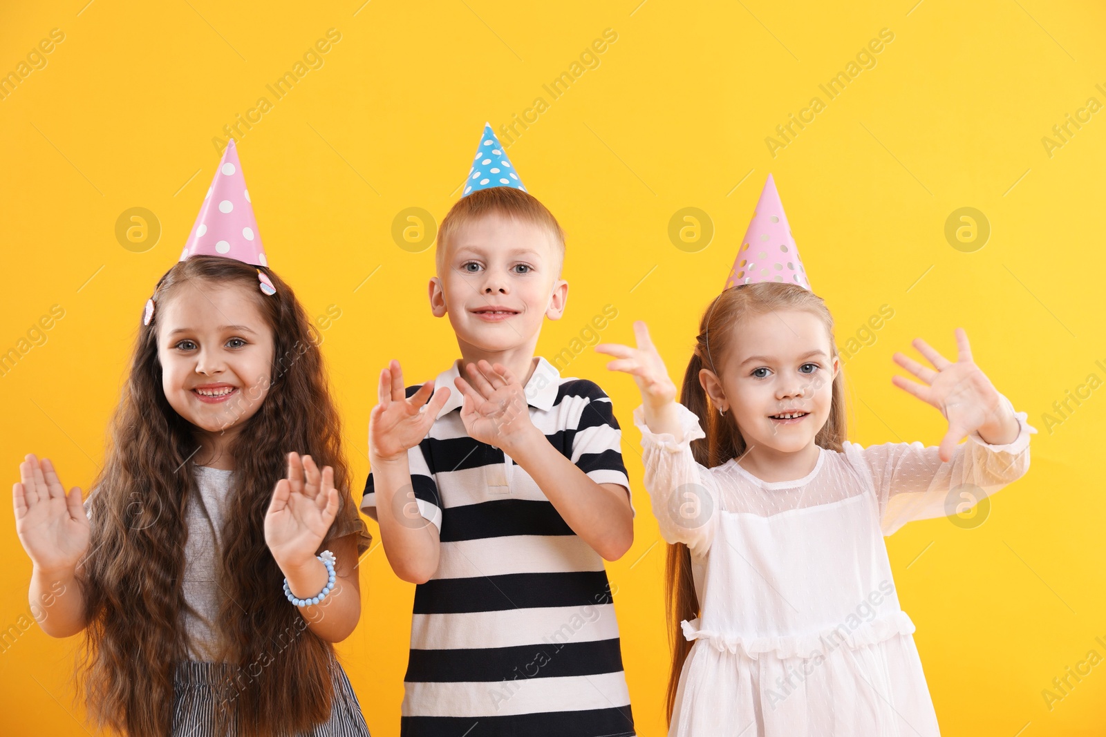 Photo of Children in conical paper hats on yellow background. Birthday surprise party