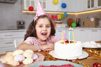 Photo of Cute little girl at table with birthday cake and different treats indoors. Surprise party