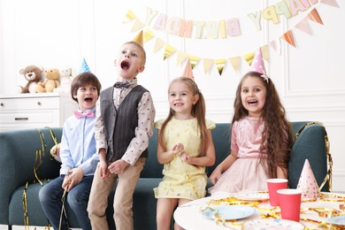 Photo of Cheerful children in conical paper hats celebrating birthday indoors. Surprise party