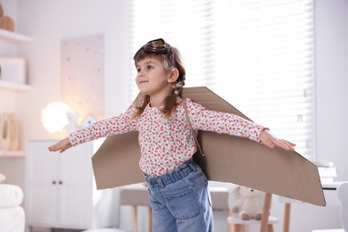 Photo of Cute little girl with cardboard plane wings and goggles playing pilot at home