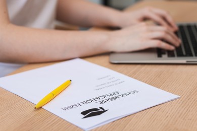 Photo of Student using laptop at wooden table, focus on scholarship application form and pen