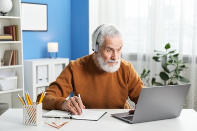 Photo of Senior man taking notes while learning online at desk indoors. Self-study