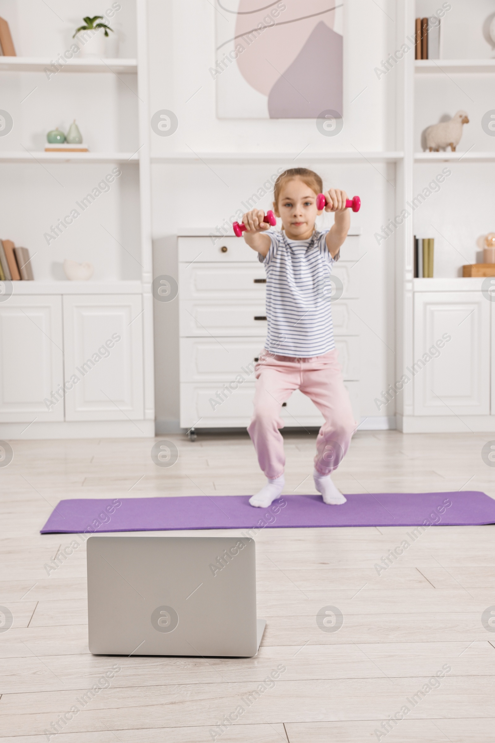 Photo of Little girl exercising with dumbbells near laptop at home. Morning routine