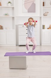 Photo of Little girl exercising with dumbbells near laptop at home. Morning routine