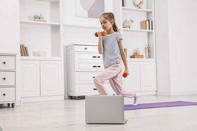 Photo of Little girl exercising with dumbbells near laptop at home, low angle view. Morning routine