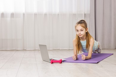 Photo of Little girl doing plank exercise near laptop at home, space for text. Morning routine