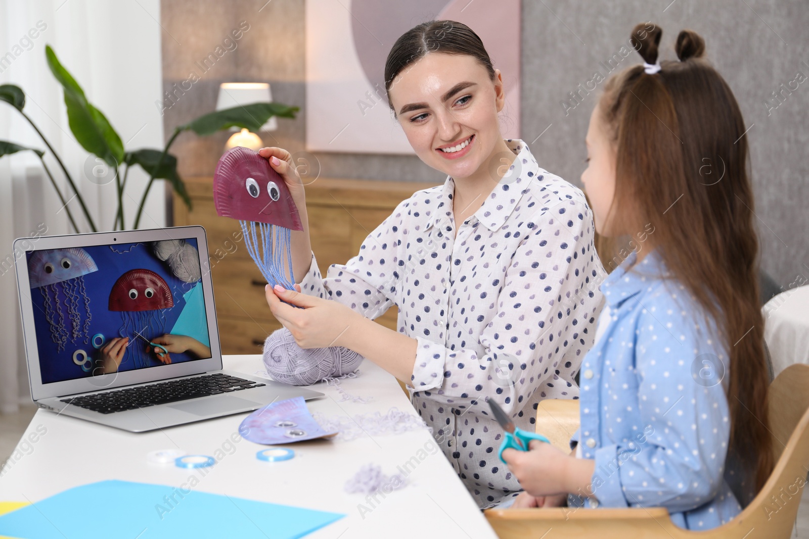 Photo of Woman and little girl making handmade jellyfish by craft tutorial video at home. Child creativity