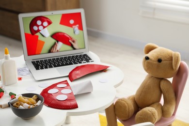 Photo of Handmade fly agaric, teddy bear and laptop with craft tutorial video on white table indoors. Child creativity