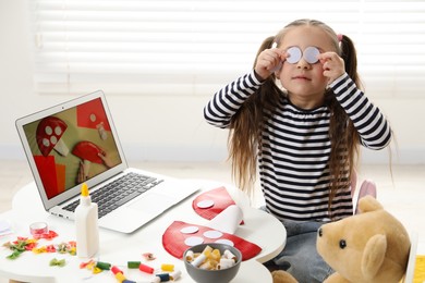 Photo of Little girl covering eyes with paper circles at white table indoors. Child creativity and craft