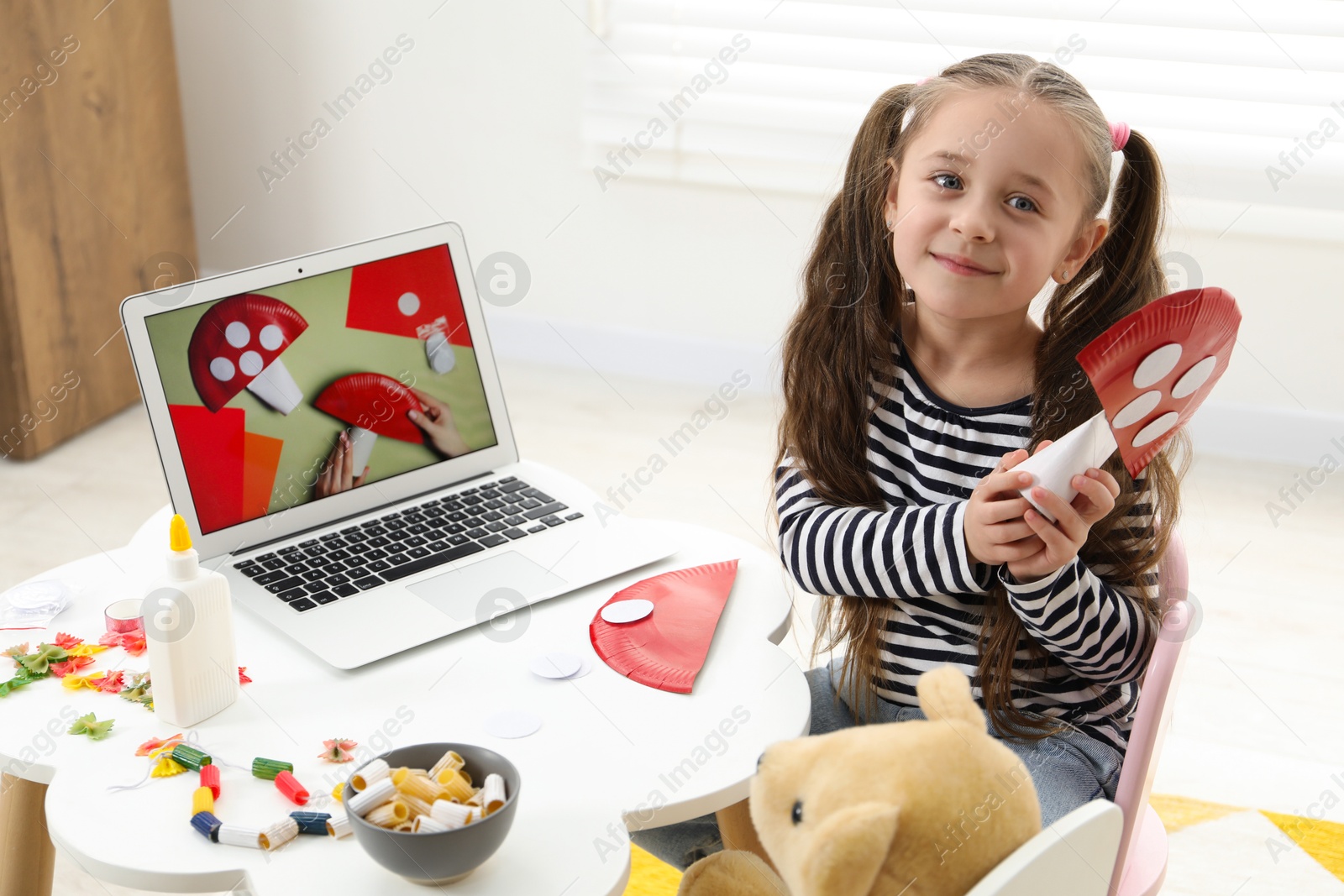 Photo of Little girl with paper fly agaric at white table indoors. Child creativity and craft