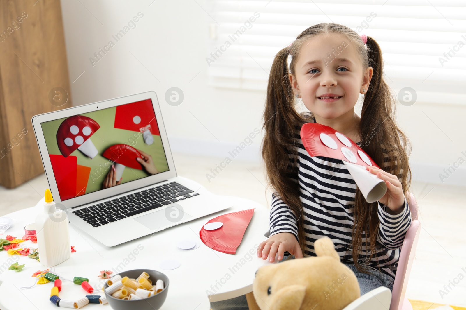 Photo of Little girl with paper fly agaric at white table indoors. Child creativity and craft