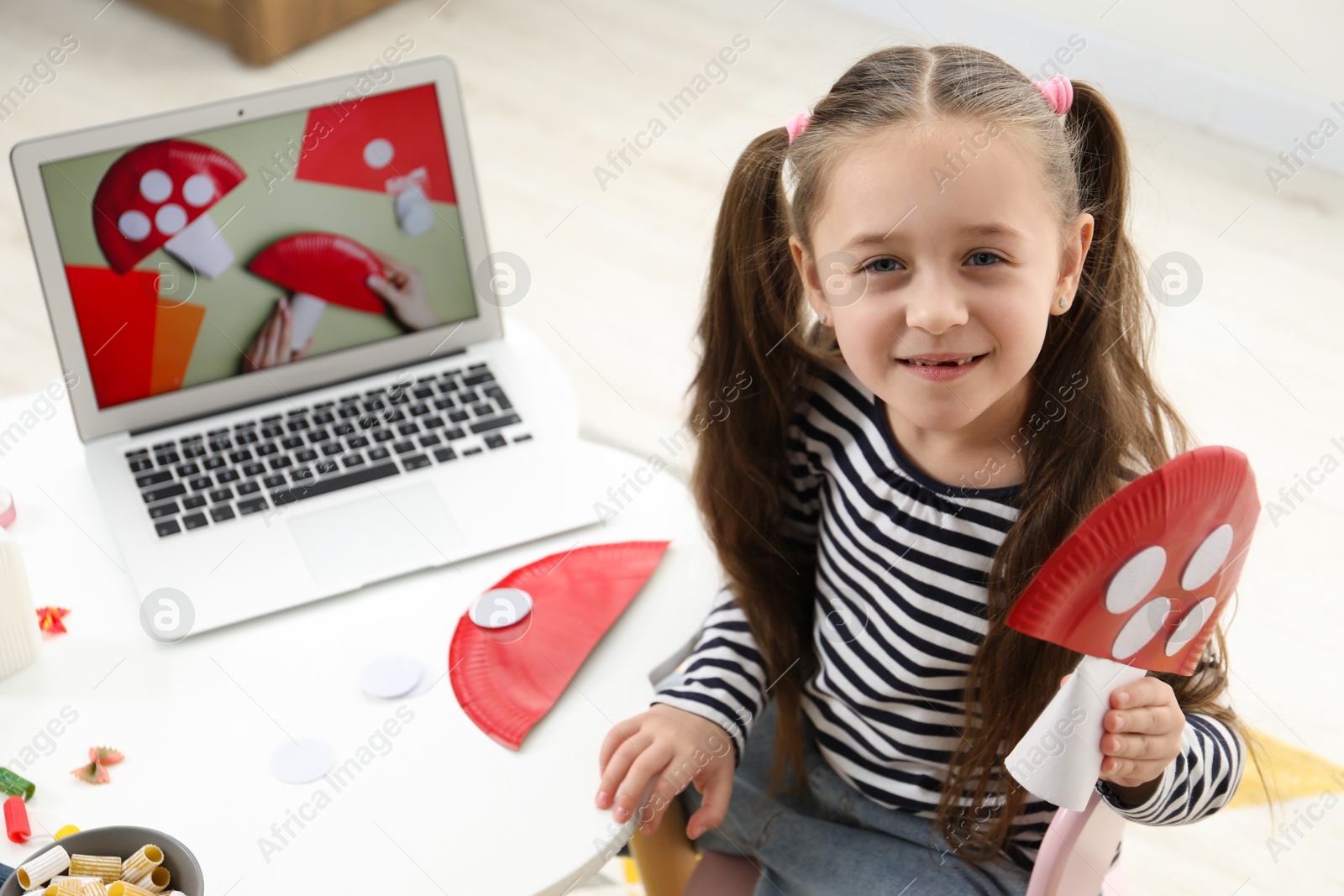 Photo of Little girl with paper fly agaric at white table indoors. Child creativity and craft