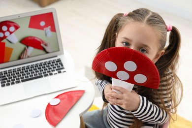 Photo of Little girl with paper fly agaric at white table indoors. Child creativity and craft