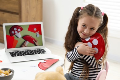 Photo of Little girl with paper fly agaric at white table indoors. Child creativity and craft