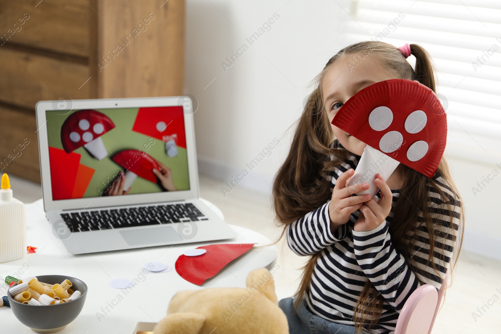 Photo of Little girl with paper fly agaric at white table indoors. Child creativity and craft