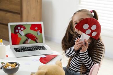 Photo of Little girl with paper fly agaric at white table indoors. Child creativity and craft