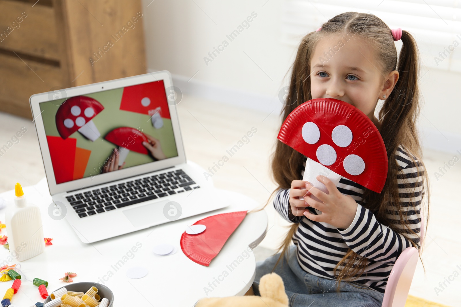 Photo of Little girl with paper fly agaric at white table indoors. Child creativity and craft