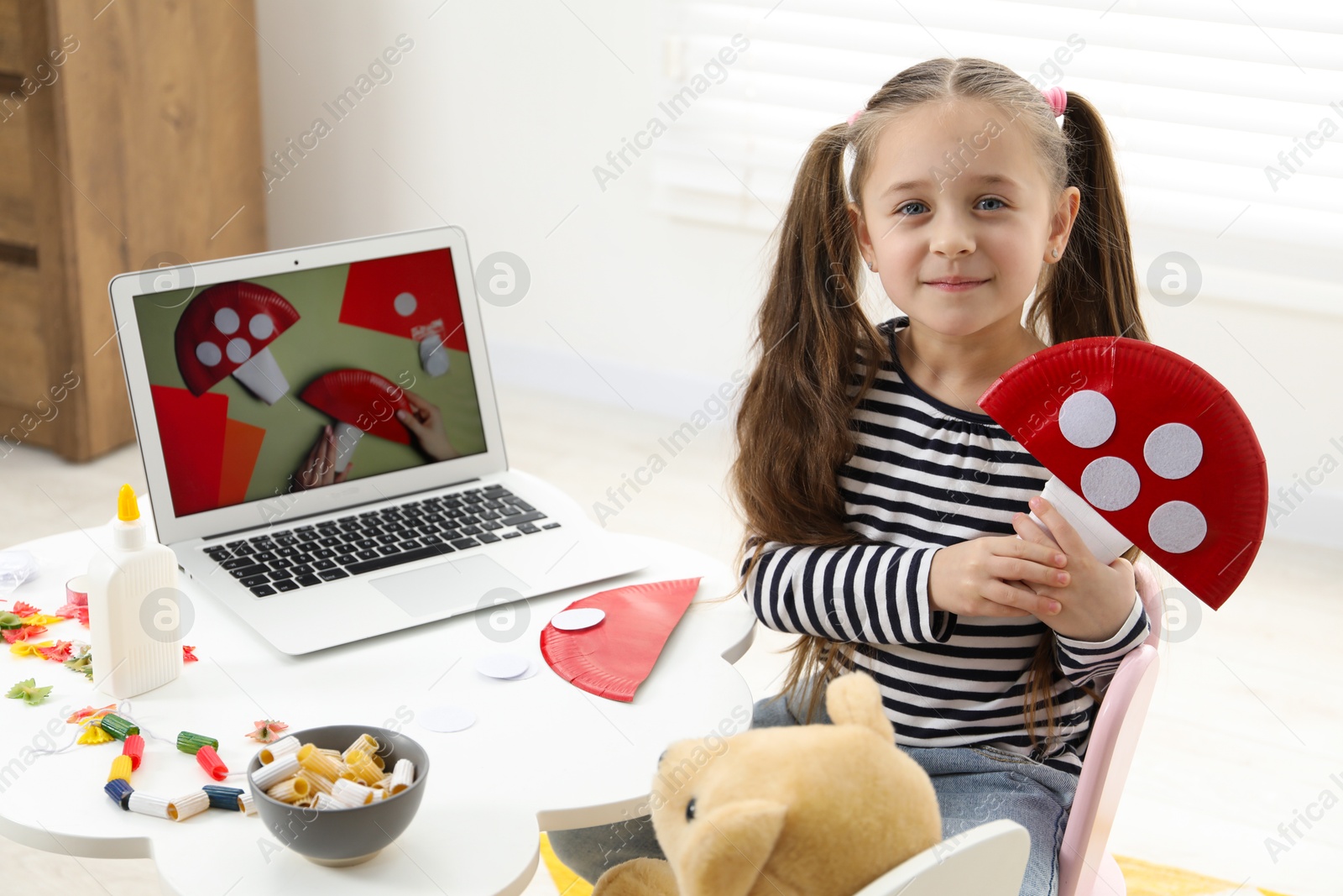 Photo of Little girl with paper fly agaric at white table indoors. Child creativity and craft