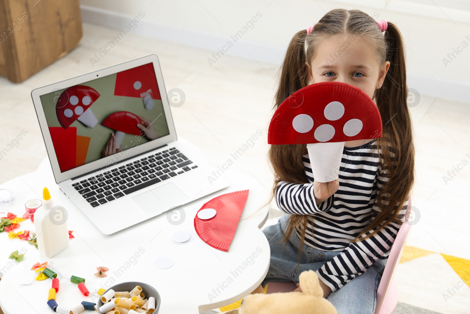 Photo of Little girl with paper fly agaric at white table indoors. Child creativity and craft