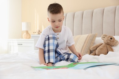 Photo of Bedtime. Cute boy with teddy bear reading book on bed indoors