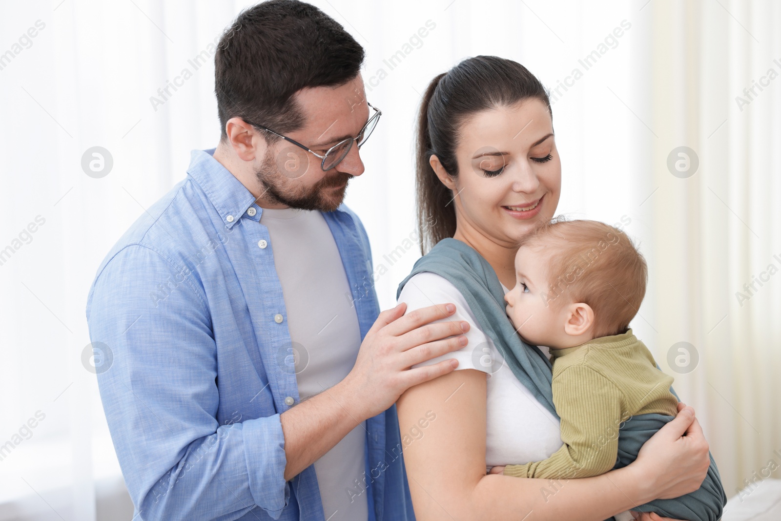 Photo of Portrait of happy family indoors. Mother holding her child in sling (baby carrier) at home