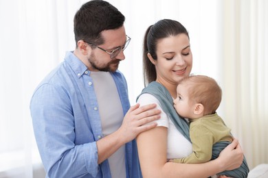 Photo of Portrait of happy family indoors. Mother holding her child in sling (baby carrier) at home