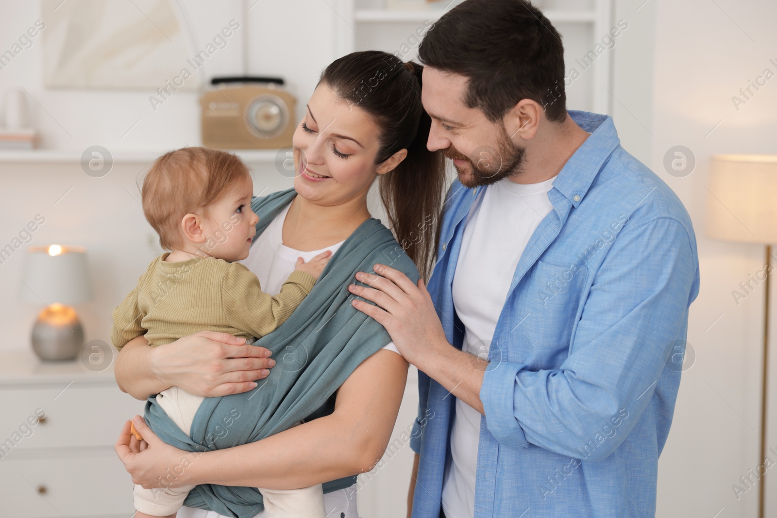 Photo of Portrait of happy family indoors. Mother holding her child in sling (baby carrier) at home