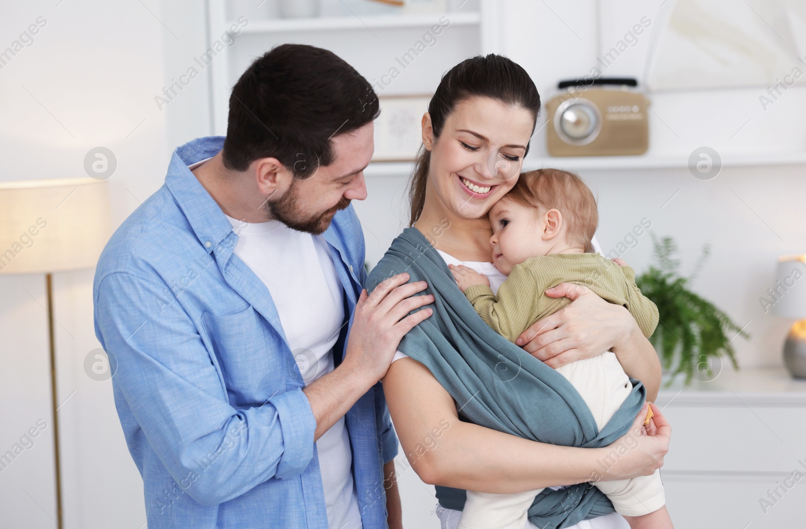 Photo of Portrait of happy family indoors. Mother holding her child in sling (baby carrier) at home