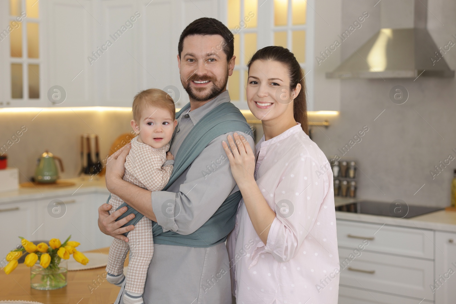 Photo of Portrait of happy family indoors. Father holding his child in sling (baby carrier) at home