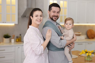 Photo of Portrait of happy family indoors. Father holding his child in sling (baby carrier) at home