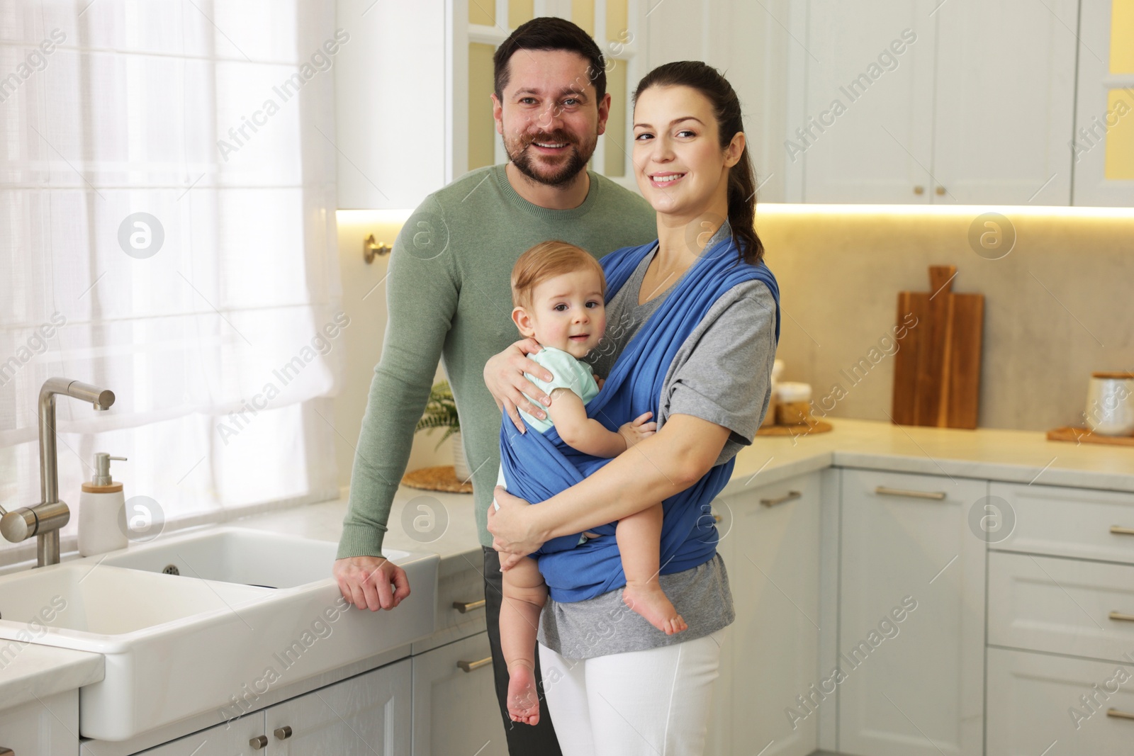 Photo of Portrait of happy family indoors. Mother holding her child in sling (baby carrier) at home