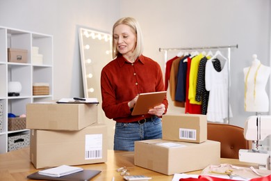 Photo of Business owner using tablet near table with parcels in her tailor shop