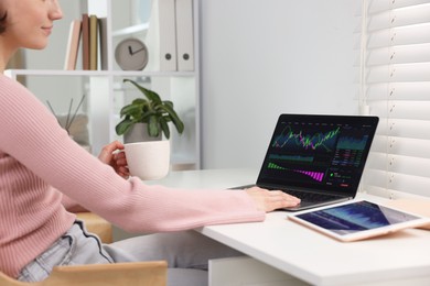 Photo of Stock exchange. Woman with cup of drink analysing financial market on laptop at white table indoors, closeup