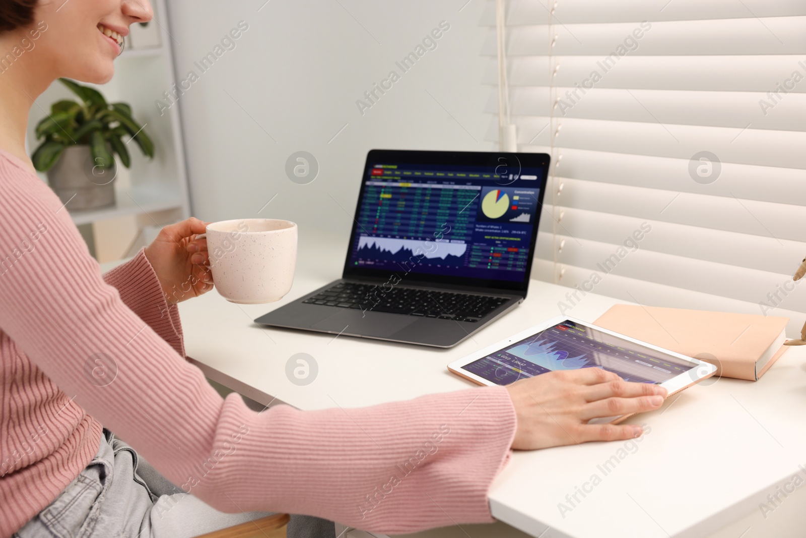 Photo of Stock exchange. Woman with cup of drink analysing financial market on tablet at table indoors, closeup