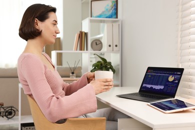 Photo of Stock exchange. Woman with cup of drink analysing financial market on laptop at white table indoors