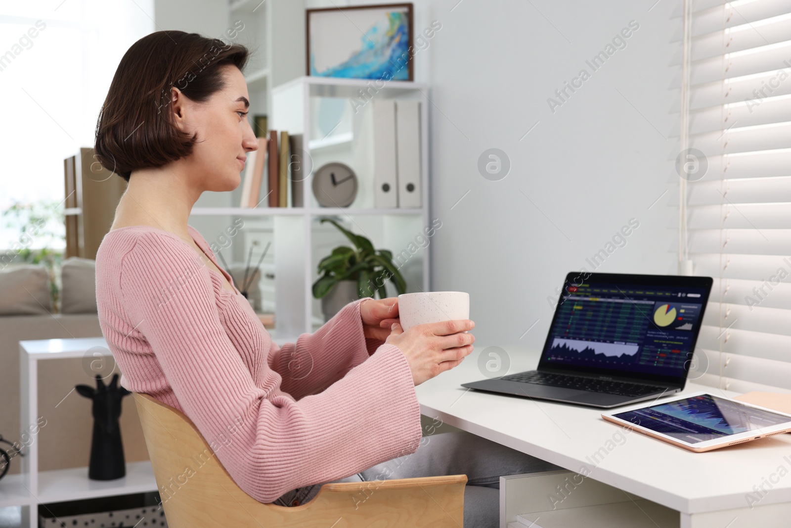 Photo of Stock exchange. Woman with cup of drink analysing financial market on laptop at white table indoors