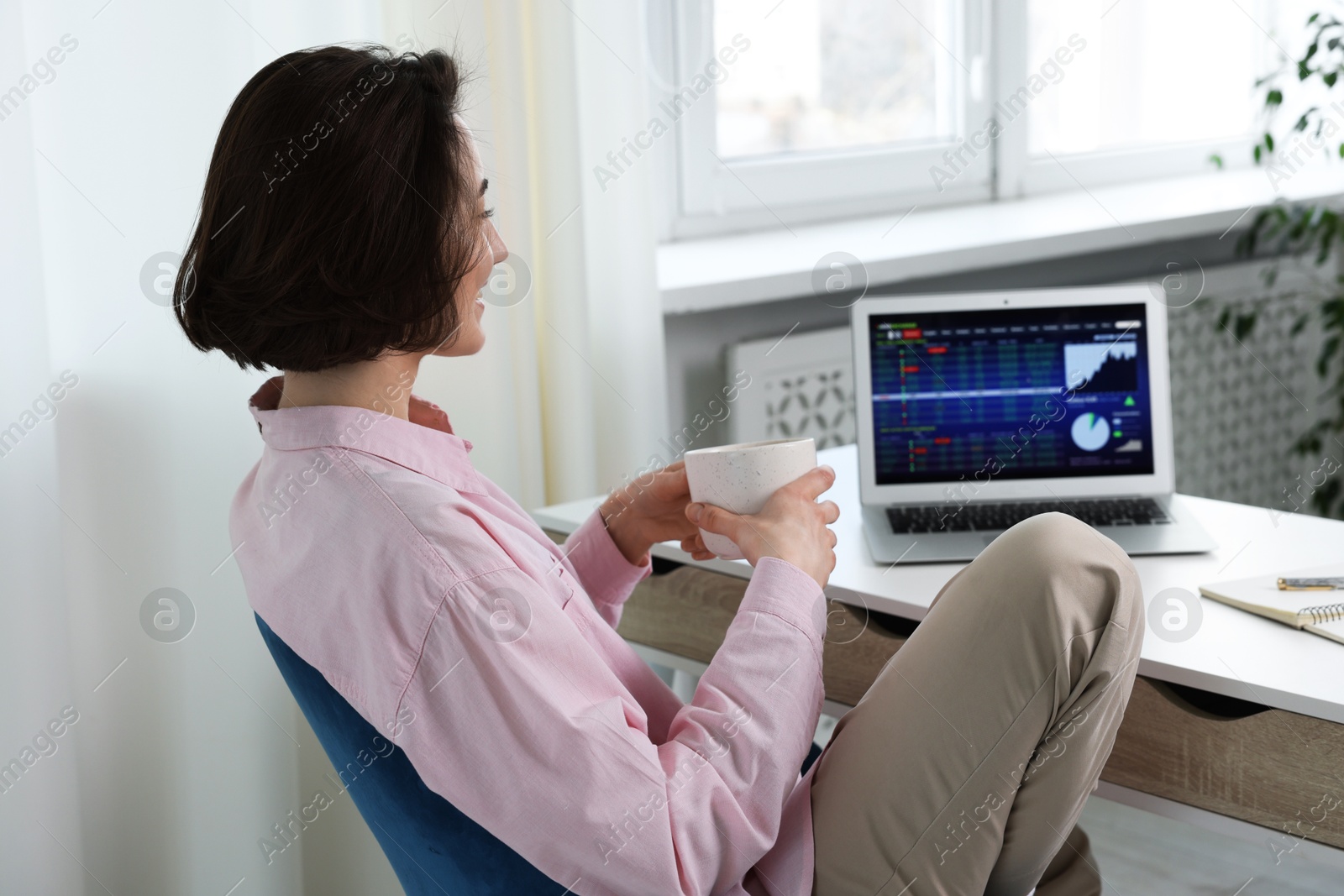 Photo of Stock exchange. Woman with cup of drink analysing financial market on laptop at white table indoors