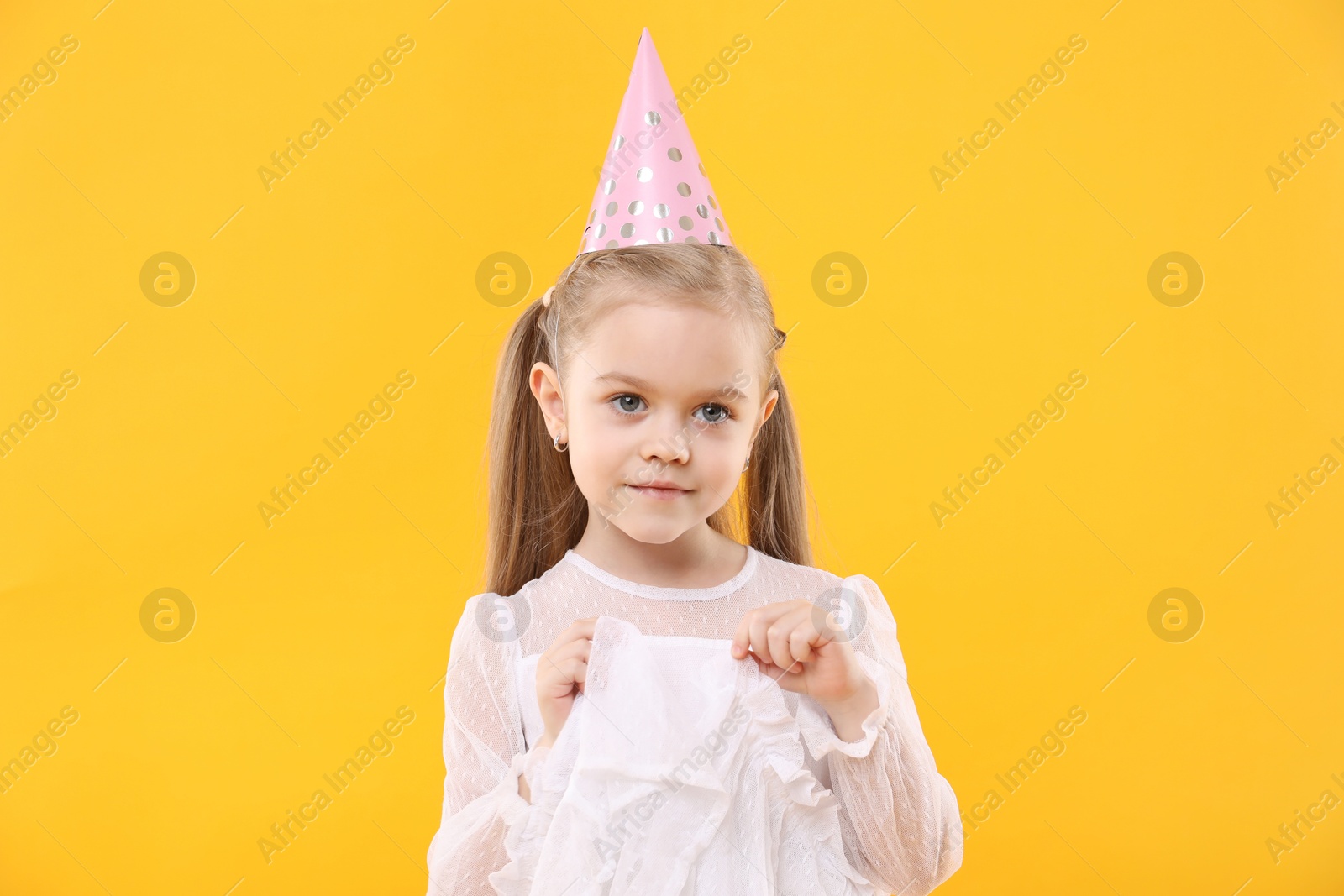 Photo of Cute little girl in conical paper hat on yellow background. Birthday surprise party