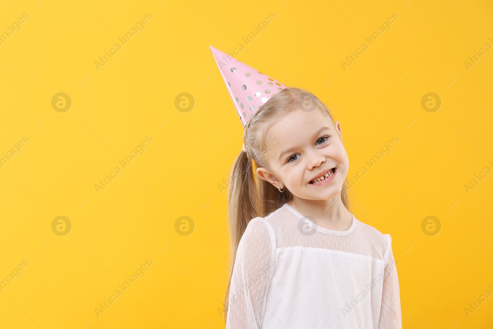 Photo of Cute little girl in conical paper hat on yellow background, space for text. Birthday surprise party